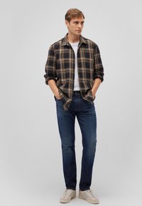 Schwarzes kariertes Flanellhemd mit orangefarbenen Akzenten, über einem weißen T-Shirt getragen, kombiniert mit blauen Jeans und beigen Sneakers.