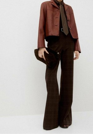 Femme portant une veste courte marron, une cravate à motifs, un pantalon taille haute à jambes larges, tenant une pochette marron foncé sur un fond uni.