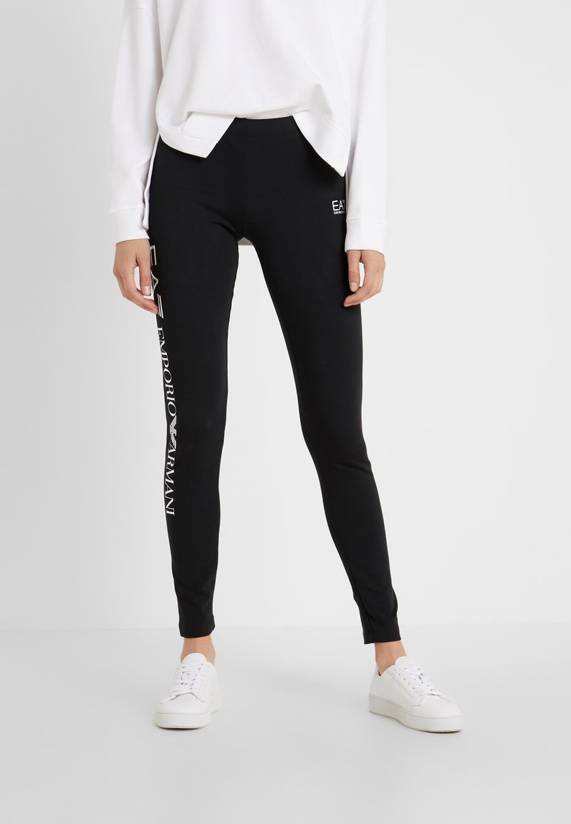 Personne portant des leggings noirs Emporio Armani avec un logo blanc sur le côté, un sweat-shirt blanc et des baskets blanches, se tenant sur un fond uni.