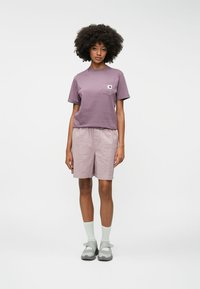 Μωβ βαμβακερό T-shirt με τσέπη στο στήθος, συνδυασμένο με ανοιχτά ροζ υφασμάτινα σορτς. Το μοντέλο φοράει γκρι σανδάλια και ανοιχτά πράσινες κάλτσες.