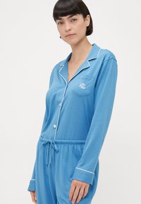 Μπλε πιτζάμα με μακριά μανίκια φτιαγμένη από μαλακό ύφασμα, με άσπρες επενδύσεις, τσέπη στο στήθος με κεντημένο λογότυπο και ελαστική μέση.