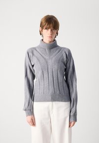 Sivi pleten sweater z visokim rebrastim ovratnikom, ki ima chevron vzorec ter rebraste manšete in spodnji del. Tkanina ima mehak občutek.