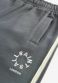 Pelēki sporta bikses ar teksturētu apdari, kas aprīkotas ar divām sānu kabatām un izšūtu logotipu "SAINT LONDON" baltā krāsā, papildinātas ar gaišu apdari.