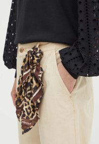 Camicia nera con dettagli in occhio di pernice, pantaloni beige con una sciarpa a volant in stampa leopardata legata in vita con accenti marroni.