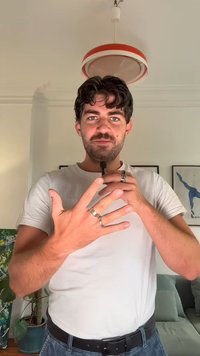 Stříbrné prsteny na prstech, bílé tričko a tmavě modré džíny. Pozadí tvoří zelený obraz a světle zbarvená pohovka.