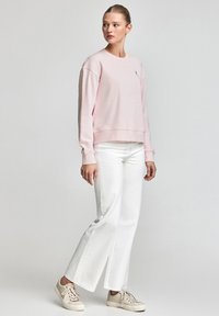 Pink sweatshirt with a crew neck and ribbed cuffs, paired with white flared jeans and beige sneakers. Minimalist design and subtle branding.