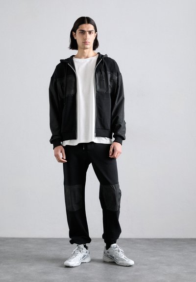 Schwarze Kapuzenjacke mit Reißverschluss, kombiniert mit einem weißen T-Shirt und schwarzen Jogginghosen mit strukturierten Kniepatches. Graue Sportschuhe.