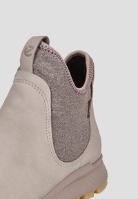 Bottine beige clair avec un panneau latéral en textile gris texturé, un col élastiqué et une semelle en caoutchouc avec un léger talon. Couture contrastante discrète.
