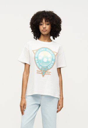 Νεαρή γυναίκα με σγουρά μαλλιά, που φοράει λευκό μπλουζάκι με μπλε και πορτοκαλί λογότυπο surf club και ανοιχτό μπλε τζιν, στέκεται μπροστά σε απλό φόντο.