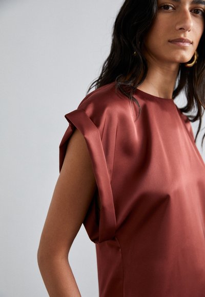 Blusa de satén burdeos con un corte relajado, mangas enrolladas y una textura suave. Las aberturas laterales mejoran el movimiento y la transpirabilidad.