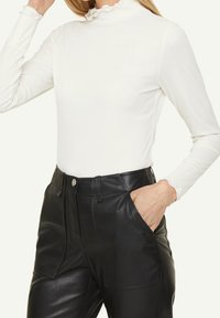 Vit ribbad långärmad topp med hög halsringning och volangdetaljer, i kombination med svarta högmidjade byxor i konstläder.