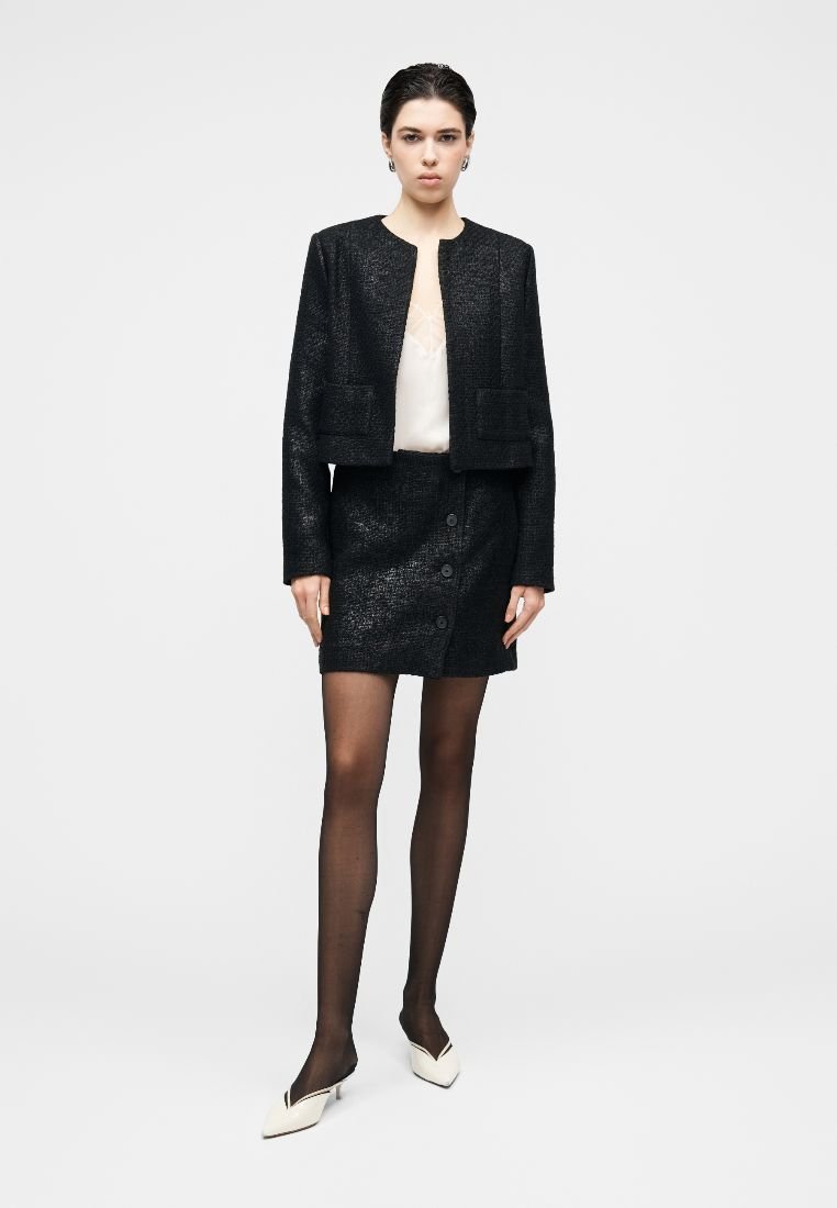 Veste noire texturée avec deux poches avant, associée à une jupe assortie avec des boutons. Portée sur un haut de couleur claire.