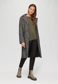 Langer Mantel mit Fischgrätenmuster in Schwarz und Weiß, mit Kragen, offener Front und tiefen Taschen. Getragen über einem grünen Oberteil und schwarzen Hosen.