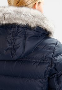 Doudoune marine avec capuche, dotée d'un col en fausse fourrure gris clair amovible. La texture est lisse et brillante, avec des coutures horizontales.