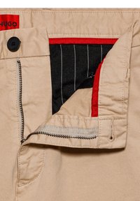 Pantalon beige avec bouton et fermeture éclair noirs, étiquette rouge indiquant "HUGO", et doublure intérieure noire avec des rayures verticales blanches et une bordure rouge.