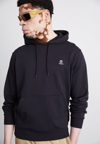 Hanorac negru din amestec de bumbac, cu un buzunar tip cangur și manșoane cu nervuri. Pata notabilă cu logo-ul Timberland pe față.