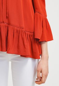 Blouse rouge texturée avec des manches volantes et un ourlet froncé. Une bordure en dentelle décorative court le long du buste, associée à un pantalon blanc.