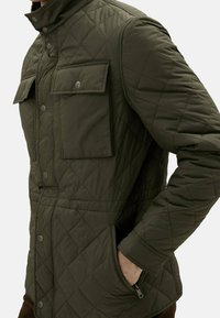 Veste verte matelassée, dotée d'un col haut, d'une fermeture à boutons-pression, de deux poches poitrine et de poches latérales avec une texture de tissu lisse et durable.