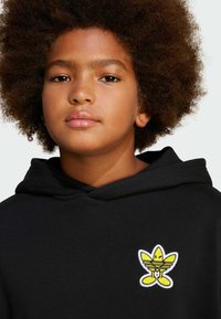 Černá mikina s kapucí a měkkou strukturou, na prsou s žlutou vyšitou logem. Mikina má stahovací šňůrku na krku.