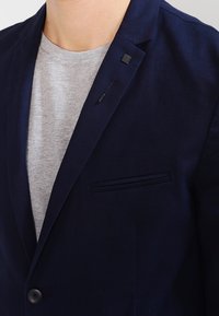 Navy blazer with a tailored fit, notch lapels, single button closing, and a gray crew-neck t-shirt underneath. Features a chest pocket and textured fabric.