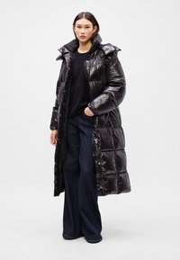 Geacă puf din material lucios, neagră, cu guler înalt, nasturi tip clapă și un design lung, matlasat. Pur tată peste un pulover negru și blugi evazați, de culoare albastră.