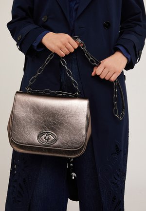 Metallisch bronze Handtasche mit strukturierter Oberfläche, ausgestattet mit einem Kettenhenkel und einem kunstvollen Emblem auf der Vorderseite.