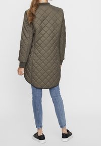 Oliivivihreä tikkikuivatut pitkä takki, jossa on pyöreä kaula-aukko ja ribatut hihat, rento malli ja kaartuva helma. Yhdistetty sinisiin farkkuihin ja mustiin lenkkareihin.