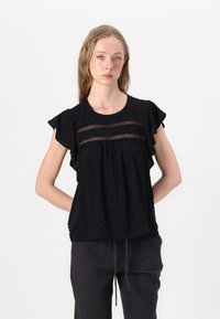 Black blouse with ruffled sleeves, textured polka-dot fabric, and mesh insets at the chest. Paired with loose dark pants.