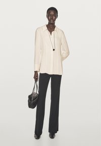 Cremefarvet bluse med krave, knaplukning foran og lange ærmer, kombineret med sorte bukser. Model bærer en sort taske med tekstur. Sorte sko.