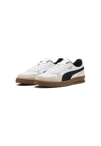 Paire de baskets basses blanches Puma avec bande latérale noire, détails en daim beige et semelles en caoutchouc marron sur fond blanc.