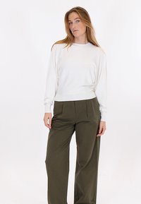 Kvinde med langt blondt hår iført en hvid langærmet bluse og løstsiddende olivengrønne bukser, stående foran en ensartet hvid baggrund.