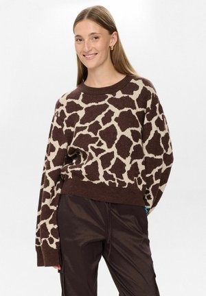 Pull marron et beige avec un motif de girafe, au design oversized avec un col rond et des manches longues. Associe à un pantalon foncé.