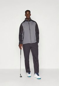 Completo da golf da uomo composto da una giacca antivento bicolore grigia e pantaloni scuri. Dotato di bastone da golf e scarpe bianche con dettagli blu.