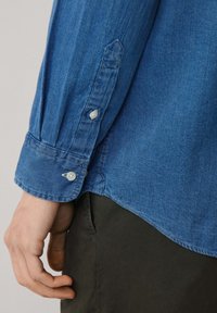 Blauwe spijkerblouse met een knoopsluiting en diagonale sluiting. Beschikt over een gestructureerde afwerking en is gecombineerd met donkergroene broek.