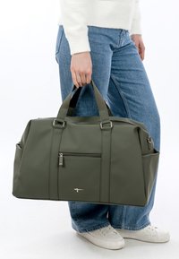 Persoon in blauwe spijkerbroek en witte sneakers die een grote olijfgroene reistas vasthoudt bij de handvatten.