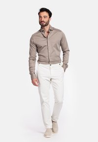 Lichtbruine overhemd met knopen, taps toelopende witte broek en beige schoenen. Het overhemd heeft een klassieke kraag en een gladde textuur.