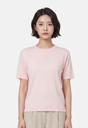 Jeune femme aux cheveux courts et foncés portant une chemise rose clair à manches courtes et un pantalon beige, face à l'avant sur un fond uni.
