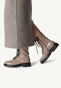 Botas de tobillo de charol beige claro con cordones negros, suelas de goma gruesa y cremalleras laterales, combinadas con una falda de lana gris.