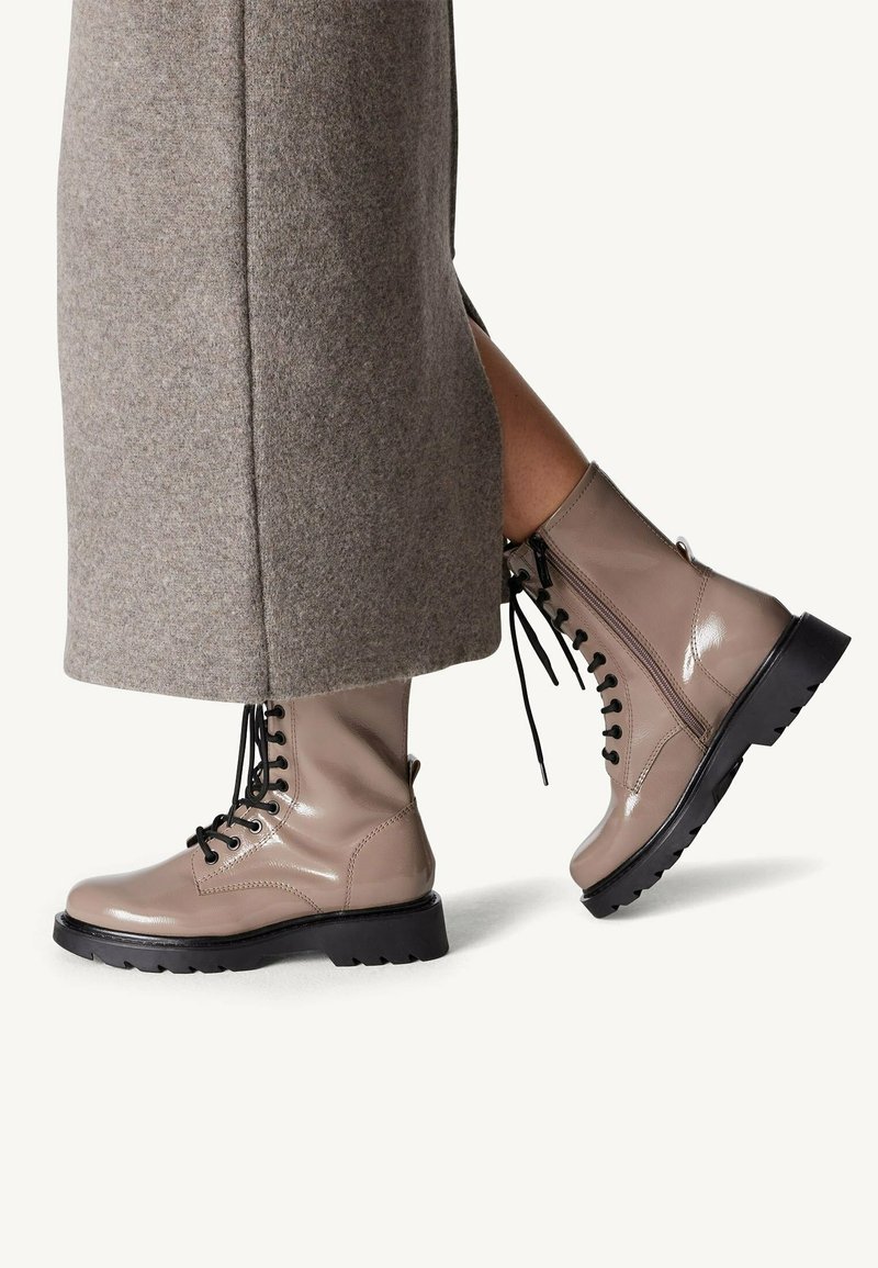 Botas de tobillo de charol beige claro con cordones negros, suelas de goma gruesa y cremalleras laterales, combinadas con una falda de lana gris.