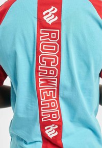 Trumpas rankovėmis marškinėliai, turintys mėlyną pagrindą su raudonomis detalėmis, didelis tekstas "ROCAWEAR", išdėstytas vertikaliai baltai ant nugaros.