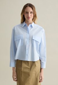 Camisa de botones de color azul claro con mangas largas, que cuenta con dos bolsillos en el pecho y un dobladillo ligeramente recortado, combinada con una falda color caqui.