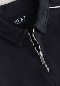 Black collared garment with silver zipper partially open, showing "NEXT Made in Bangladesh" label inside the neckline.