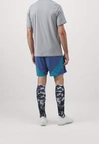 Uomo con camicia grigia, pantaloni corte blu e verde acqua, calze sportive a ginocchio nere e bianche a fantasia e scarpe bianche, in piedi di spalle contro uno sfondo semplice.