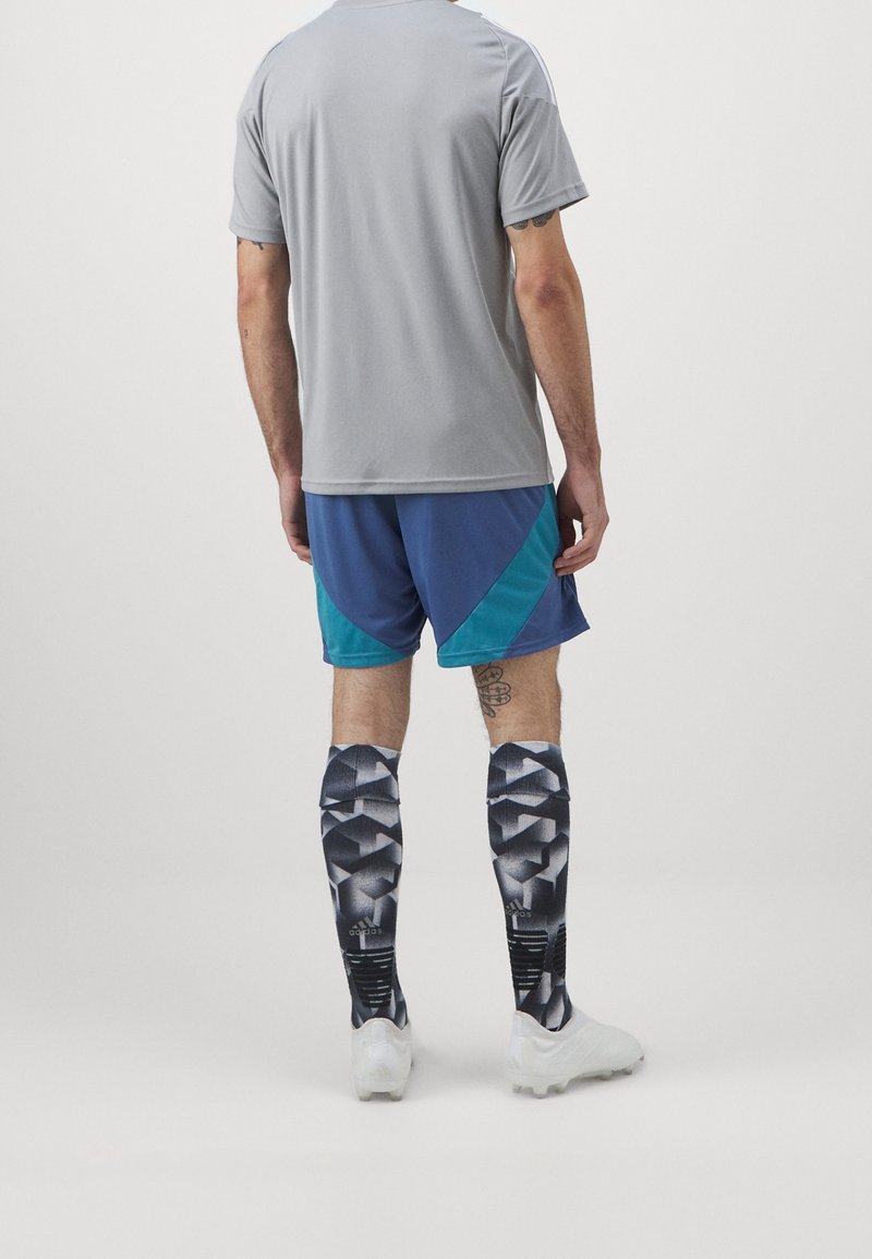 Uomo con camicia grigia, pantaloni corte blu e verde acqua, calze sportive a ginocchio nere e bianche a fantasia e scarpe bianche, in piedi di spalle contro uno sfondo semplice.
