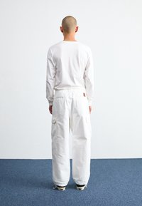 Chemise blanche à manches longues et pantalon cargo blanc avec poches latérales, porté avec des chaussures noires, vu de dos contre un mur uni.