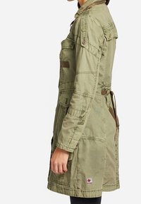 Kaki grön arbetsjacka med bälte i midjan, knäppte axelklaffar och två stora framfickor. Strukturerad tyg med sömnadsdetaljer.