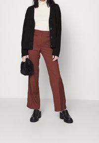 Cárdigan negro de punto con botones, jersey de cuello vuelto crema, pantalones marrones acampanados y zapatos negros de plataforma. Pequeño bolso negro texturizado en la mano.