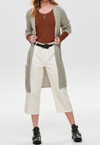 Lange, open voorkant cardigan in lichtgrijze tricot, bruin aansluitend topje, witte cropped broek en zwarte enkellaarsjes met zilveren studs.