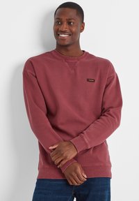 Smiling man wearing a faded red TOG24 sweatshirt and blue jeans stands with arms crossed against a plain white background.