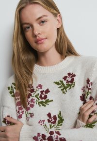 Mujer joven con cabello largo y rubio, vistiendo un suéter crema con tejido floral en tonos morados y verdes, con los brazos cruzados suavemente.